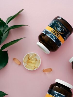 Flat lay of supplement bottles and capsules on pink with green leaves.